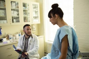 A transgender woman in a hospital gown speaking to her doctor, a transman